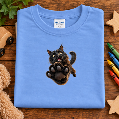 A folded carolina blue Gildan Youth cotton t-shirt with a graphic of a jumping black cat, displayed on a wooden surface with toys and crayons.