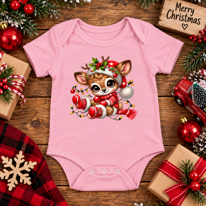 A pink short-sleeve baby bodysuit laid flat on a rustic wooden surface, featuring a large cartoon Christmas deer graphic on the front. The bodysuit is surrounded by holiday-themed props including wrapped gifts, red ornaments, pine branches, a red plaid cloth, a wooden sign reading 'Merry Christmas,' and a red toy truck.