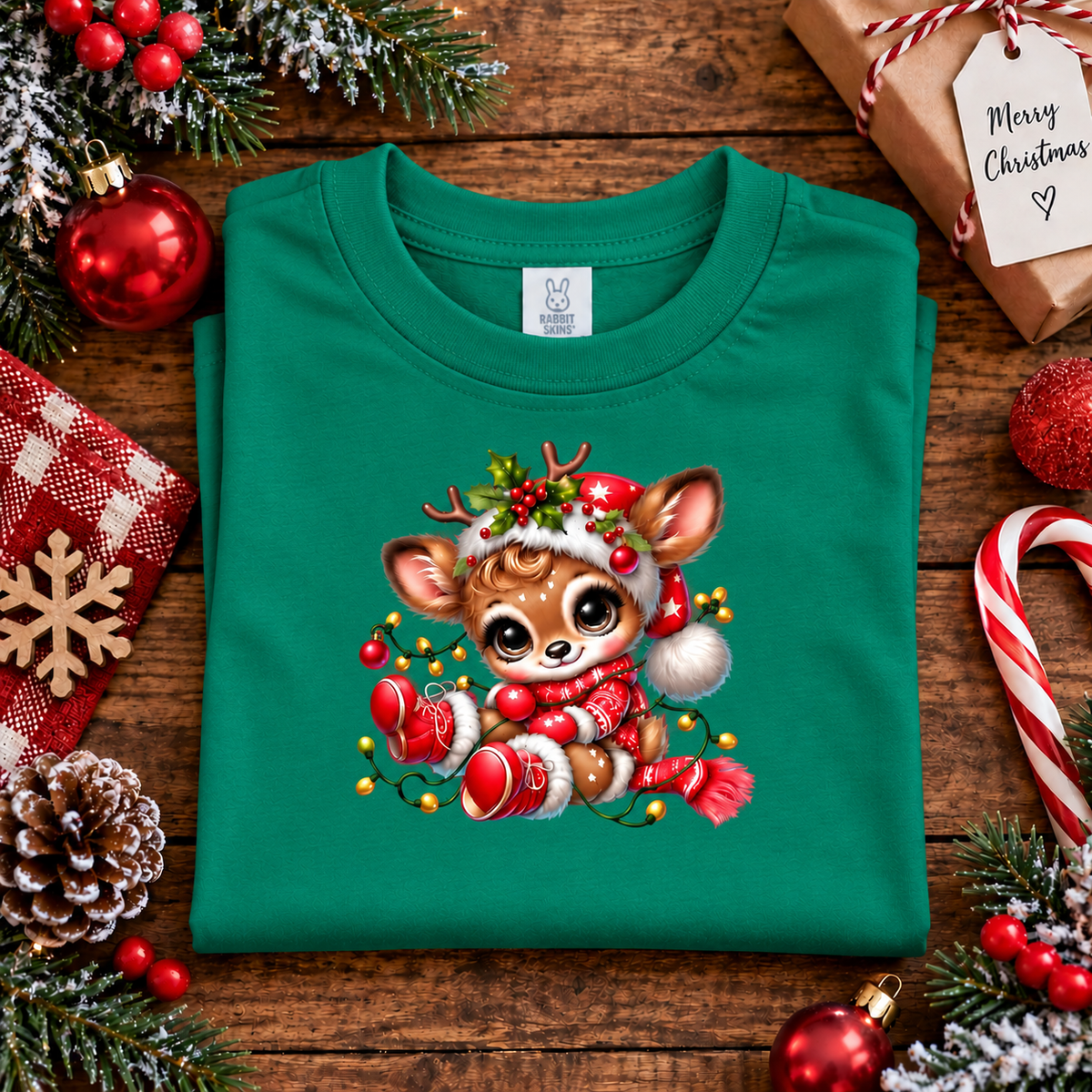 A folded green crewneck t-shirt with a cute cartoon Christmas deer graphic, styled on a rustic wooden table with holiday decorations like candy canes, ornaments, and a 'Merry Christmas' gift bag.