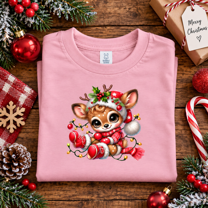 A folded light pink crewneck t-shirt with a cute cartoon Christmas deer graphic, styled on a rustic wooden background with holiday decorations including ornaments, candy canes, pinecones, and a wrapped gift.