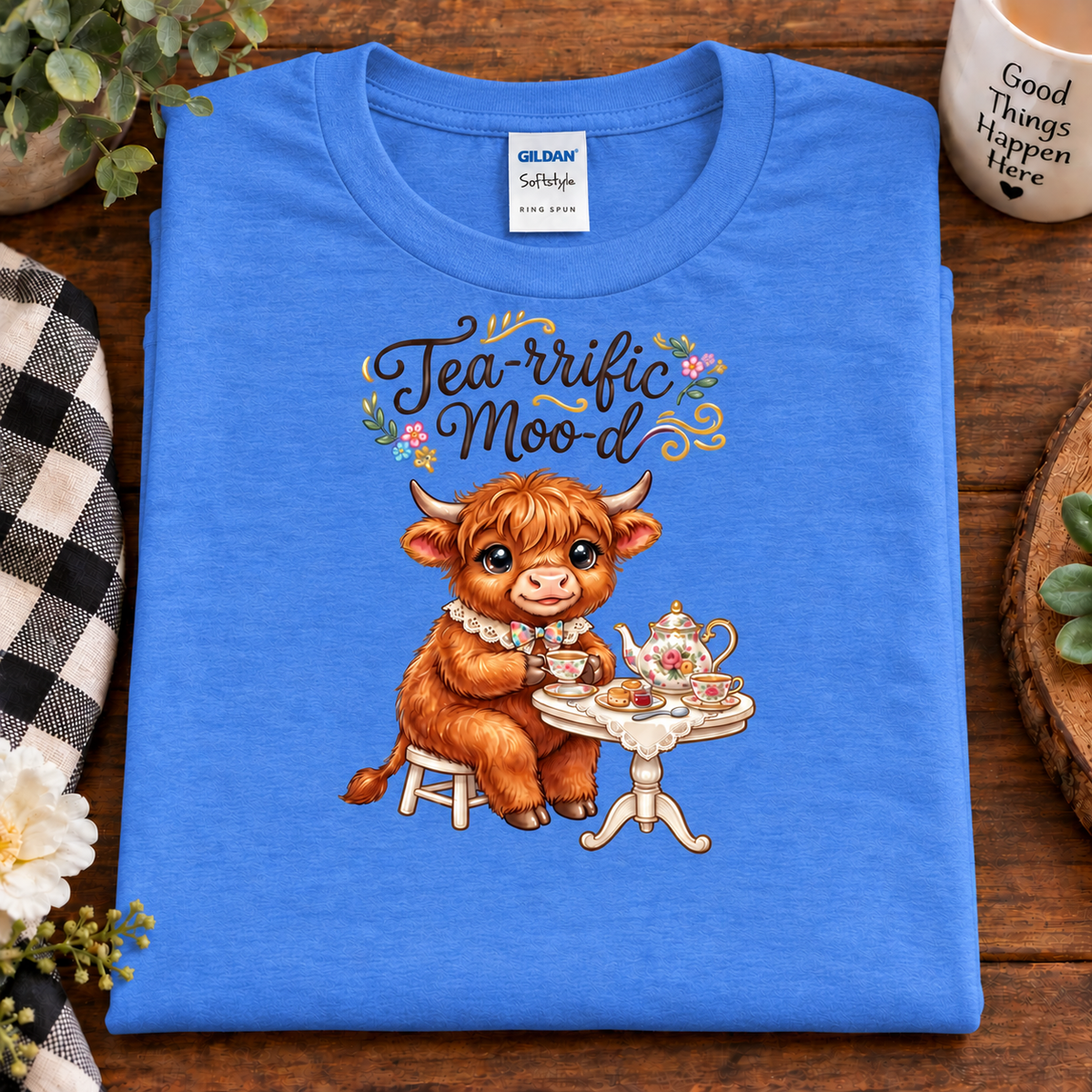 A folded heather royal Gildan Softstyle t-shirt with a graphic of a cute brown highland cow sitting at a tea table under the text 'Tea-rific Mood'. The shirt is displayed on a wooden table with lifestyle props including a mug, flowers, and a checkered cloth.