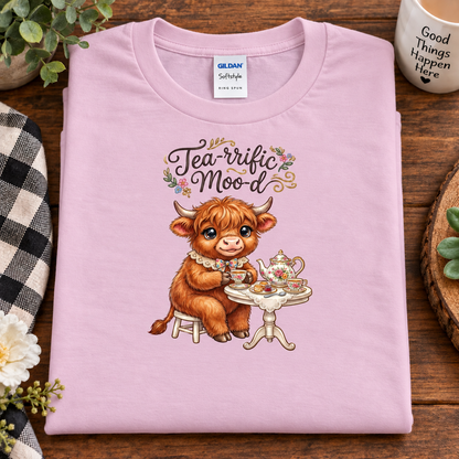 A folded light pink Gildan Softstyle T-shirt with a graphic of a brown highland cow sitting at a tea table, under the words 'Tea-rific Mood' with small flowers. The shirt is displayed on a wooden surface with lifestyle props including a mug, plant, and checkered cloth.