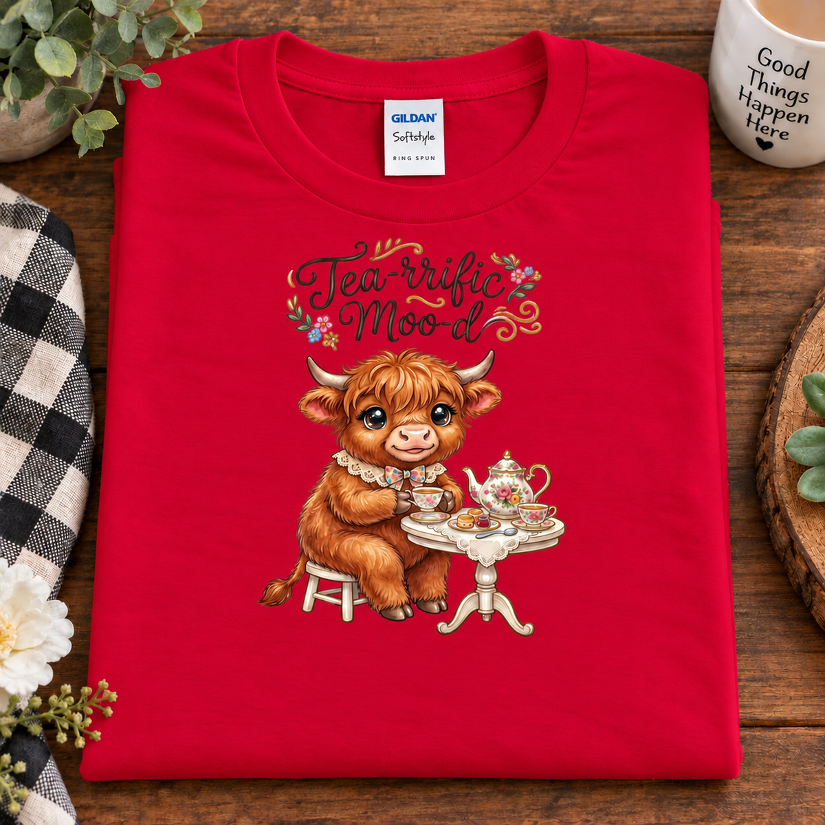A folded red Gildan Softstyle t-shirt with a graphic of a Scottish highland cow having tea, displayed on a wooden tabletop with lifestyle props.