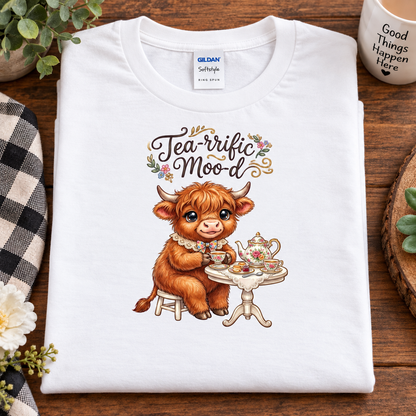 A folded white Gildan Softstyle T-shirt with a colorful graphic of a brown Scottish highland cow sitting at a tea table under the text 'Tea-rrific Mood', displayed on a wooden surface with lifestyle props.