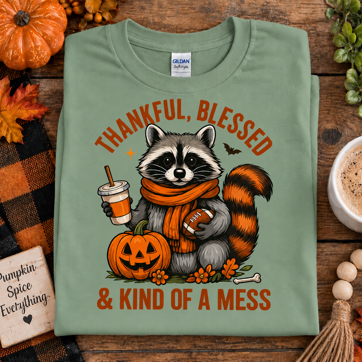 A folded sage green t-shirt with a cartoon raccoon graphic holding a football and pumpkin, surrounded by autumn props on a wooden surface.