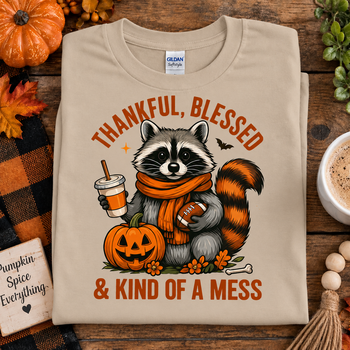 A sand colored Gildan Softstyle t-shirt is displayed flat on a rustic wooden table decorated for autumn. The shirt features a cartoon raccoon wearing an orange scarf, holding a football and a pumpkin, with the text 'THANKFUL, BLESSED & KIND OF A MESS' in orange.