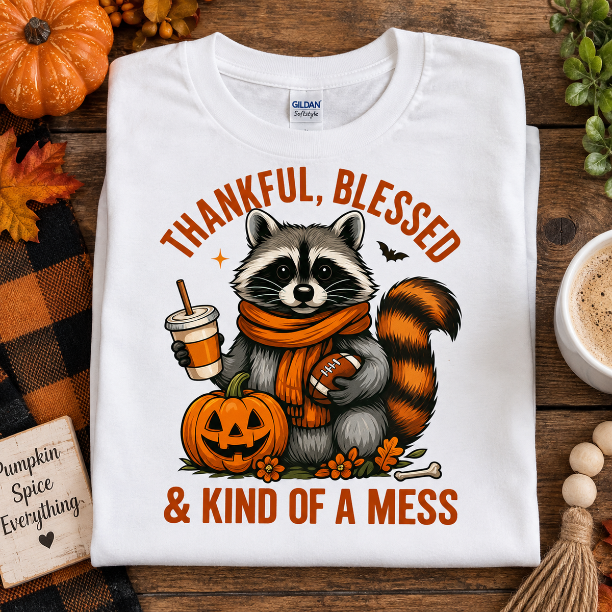 A folded white Gildan Comfort T-shirt with a humorous cartoon raccoon graphic, styled in a cozy autumn flat-lay setting with pumpkins, fall leaves, and a cup of coffee.