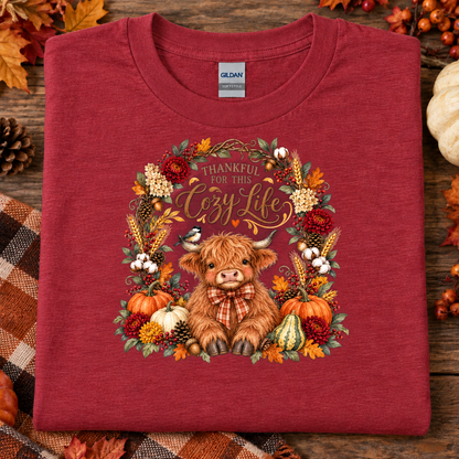 A folded heather cardinal red Gildan t-shirt with a detailed autumn-themed graphic of a shaggy brown Highland cow surrounded by pumpkins, corn, and fall foliage with the text 'THANKFUL FOR THIS Cozy Life'. The shirt is styled on a rustic wooden background with autumn leaves, a pinecone, and a plaid blanket.