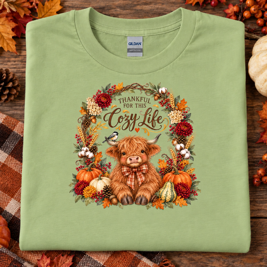 A folded pistachio colored t-shirt with a cozy autumn graphic featuring a highland cow, pumpkins, and the text 'THANKFUL FOR THIS Cozy Life'. The shirt is displayed flat on a rustic wooden background decorated with fall leaves, pinecones, and a plaid blanket.