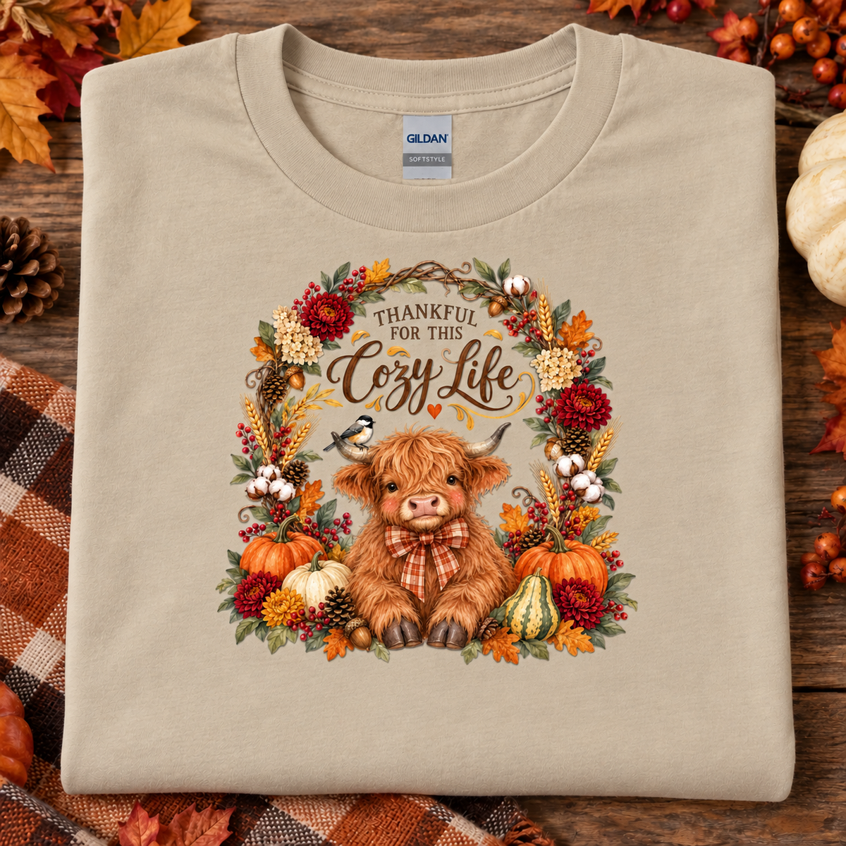 A folded sand colored Gildan t-shirt with a cozy autumn-themed graphic print featuring a Highland cow, pumpkins, and the text 'THANKFUL FOR THIS Cozy Life'. The shirt is styled on a rustic wooden background with fall leaves, pinecones, and a plaid blanket.