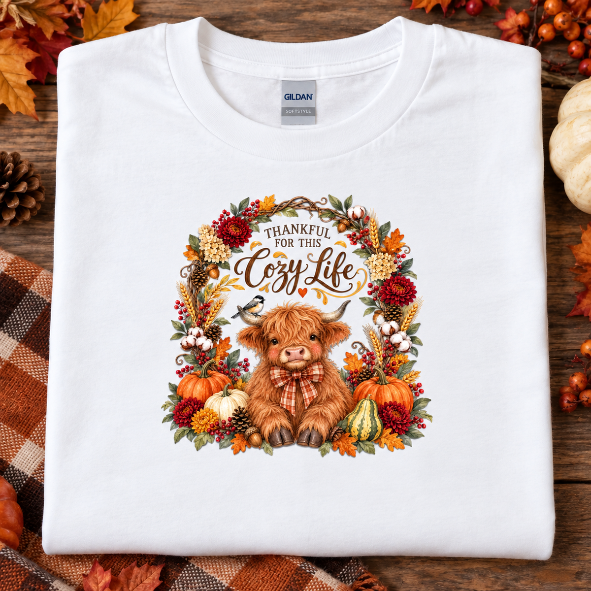 A folded white Gildan t-shirt with a cozy autumn graphic, displayed on a rustic wooden background with fall leaves, pumpkins, and a plaid blanket. The graphic features a cute brown highland cow surrounded by a wreath of autumn leaves, pumpkins, and the text 'THANKFUL FOR THE Cozy Life'.