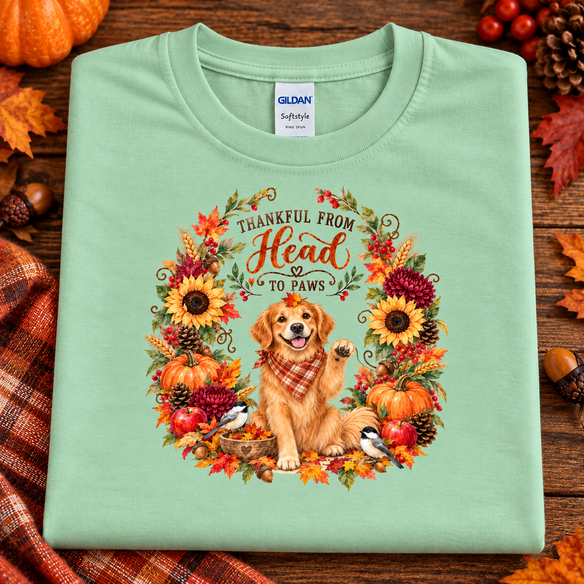 A folded mint green Gildan Softstyle T-shirt with a large autumn-themed graphic of a golden retriever inside a floral wreath and the text 'THANKFUL FROM Head TO Paws,' displayed on a rustic wood surface with fall leaves, pumpkins, and plaid fabric around it.