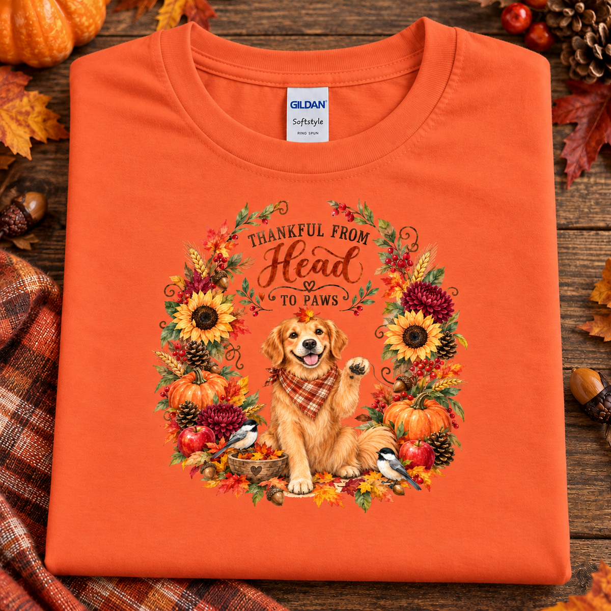 An orange folded Gildan Softstyle T-shirt with a large autumn-themed graphic on the front, featuring a golden retriever dog in a plaid scarf surrounded by sunflowers, pumpkins, birds, and the text 'THANKFUL FROM Head TO PAWS,' styled on a rustic wood surface with fall leaves and pumpkins.