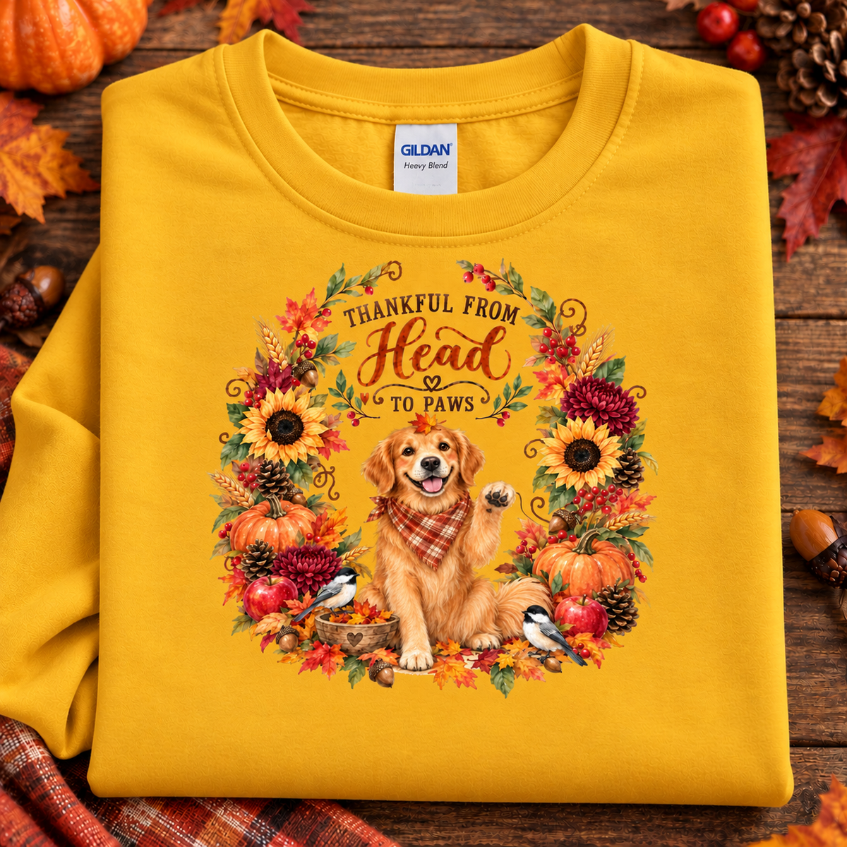 A folded gold Gildan t-shirt with a detailed autumn graphic featuring a golden retriever, pumpkins, sunflowers, and birds inside a wreath with the text 'THANKFUL FROM Head TO Paws'. The shirt is styled on a rustic wooden background with fall leaves and acorns.
