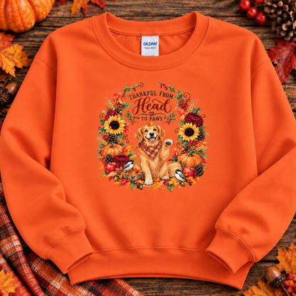 An orange crewneck sweatshirt laid flat on a rustic wooden surface with autumn props. The front features a large graphic of a golden retriever dog surrounded by sunflowers, pinecones, berries, and leaves, with the text 'THANKFUL FROM Head TO Paws' inside a floral wreath.