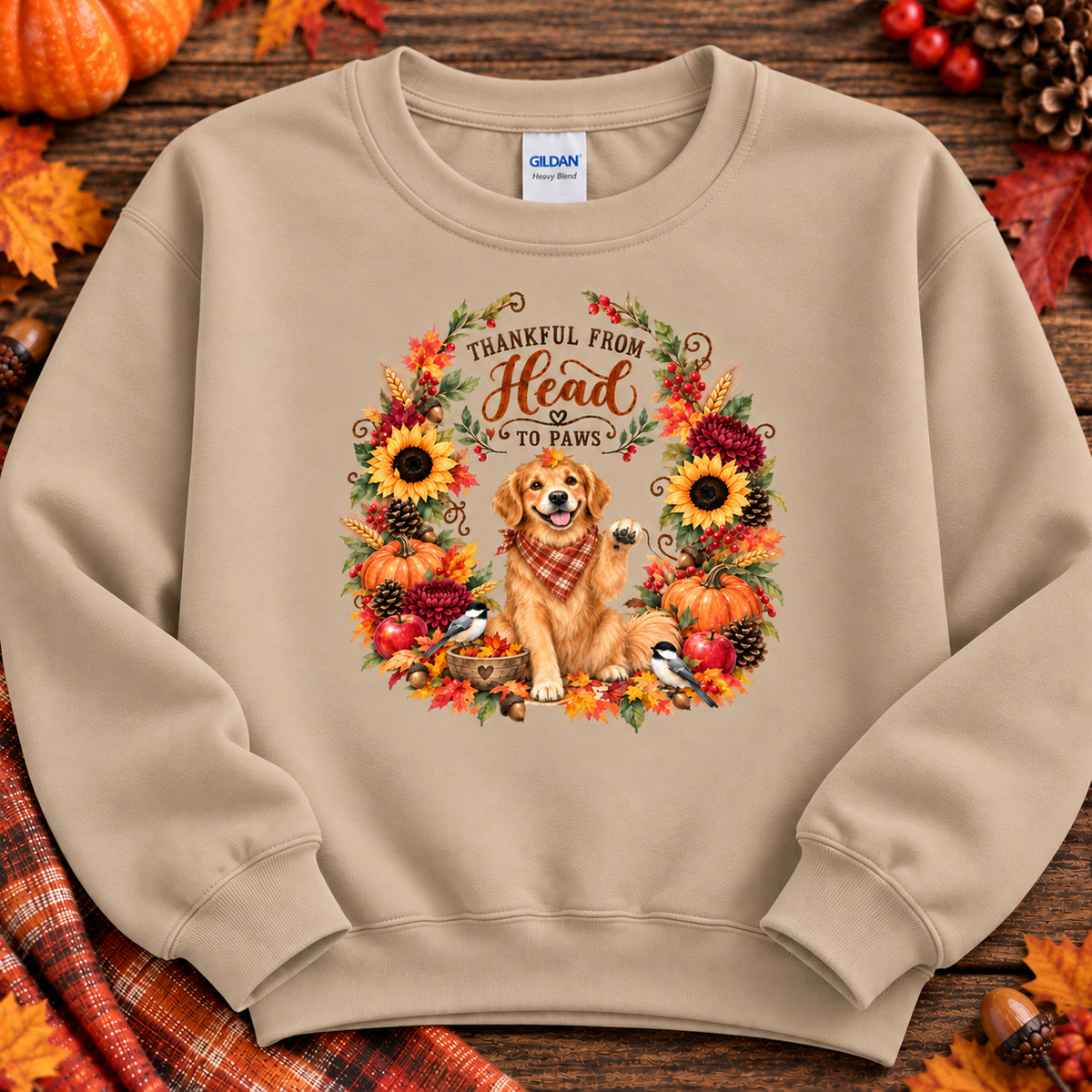A sand crewneck sweatshirt laid flat on a rustic wooden surface with autumn props. The sweatshirt features a large graphic print of a golden retriever wearing a plaid scarf, surrounded by sunflowers, pumpkins, birds, and the text 'THANKFUL FROM Head TO PAWS'.