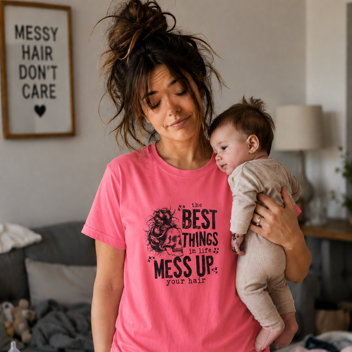 Woman wearing a coral silk colored  t-shirt with text " The best things in life Mess up your hair" holding a baby in a living room.