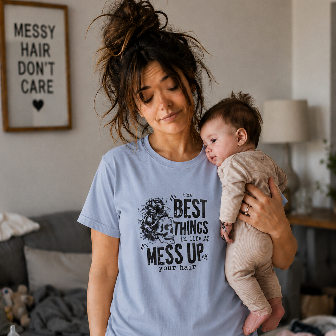 Woman wearing a light blue t-shirt with text " The best things in life Mess up your hair" holding a baby in a living room.