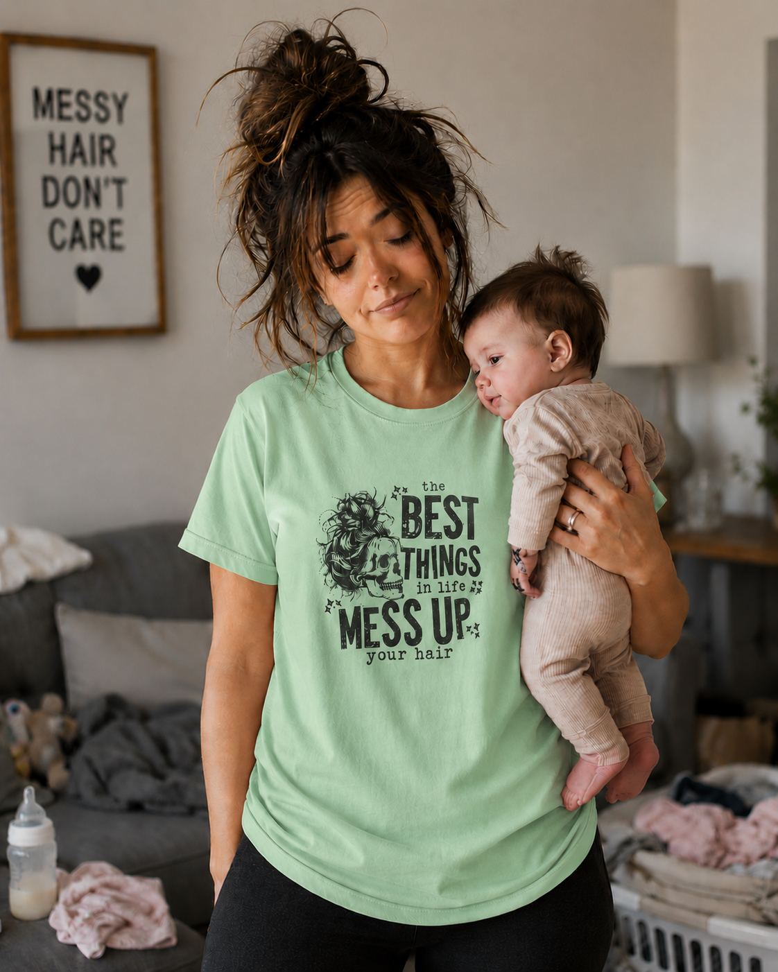 Woman wearing a mint green t-shirt with text " The best things in life Mess up your hair" holding a baby in a living room.