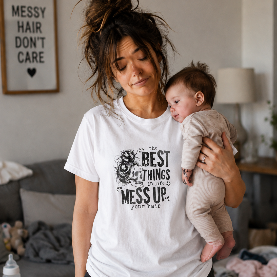 Woman wearing a white t-shirt with text " The best things in life Mess up your hair" holding a baby in a living room.
