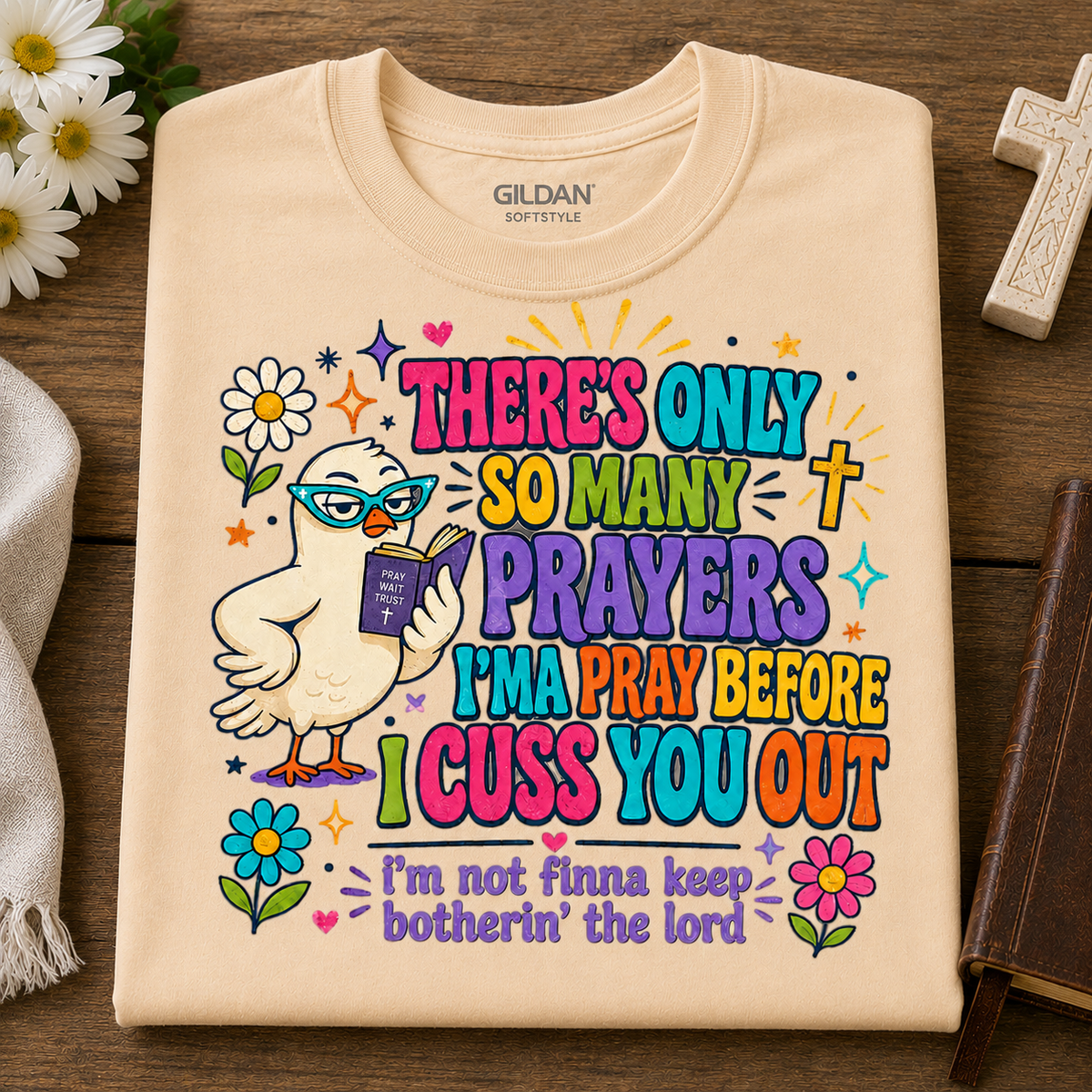 A folded natural colored Gildan Softstyle t-shirt with a colorful graphic print of a cartoon dove reading a book, surrounded by flowers and the text 'THERE'S ONLY SO MANY PRAYERS I'M PRAY BEFORE I CUSS YOU OUT' and 'I'm not finna keep botherin' the lord', displayed on a wooden background with decorative props.