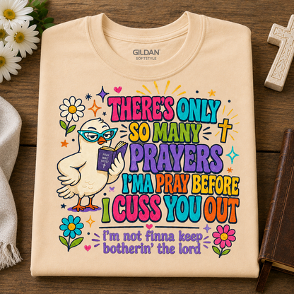 A folded natural colored Gildan Softstyle t-shirt with a colorful graphic print of a cartoon dove reading a book, surrounded by flowers and the text 'THERE'S ONLY SO MANY PRAYERS I'M PRAY BEFORE I CUSS YOU OUT' and 'I'm not finna keep botherin' the lord', displayed on a wooden background with decorative props.