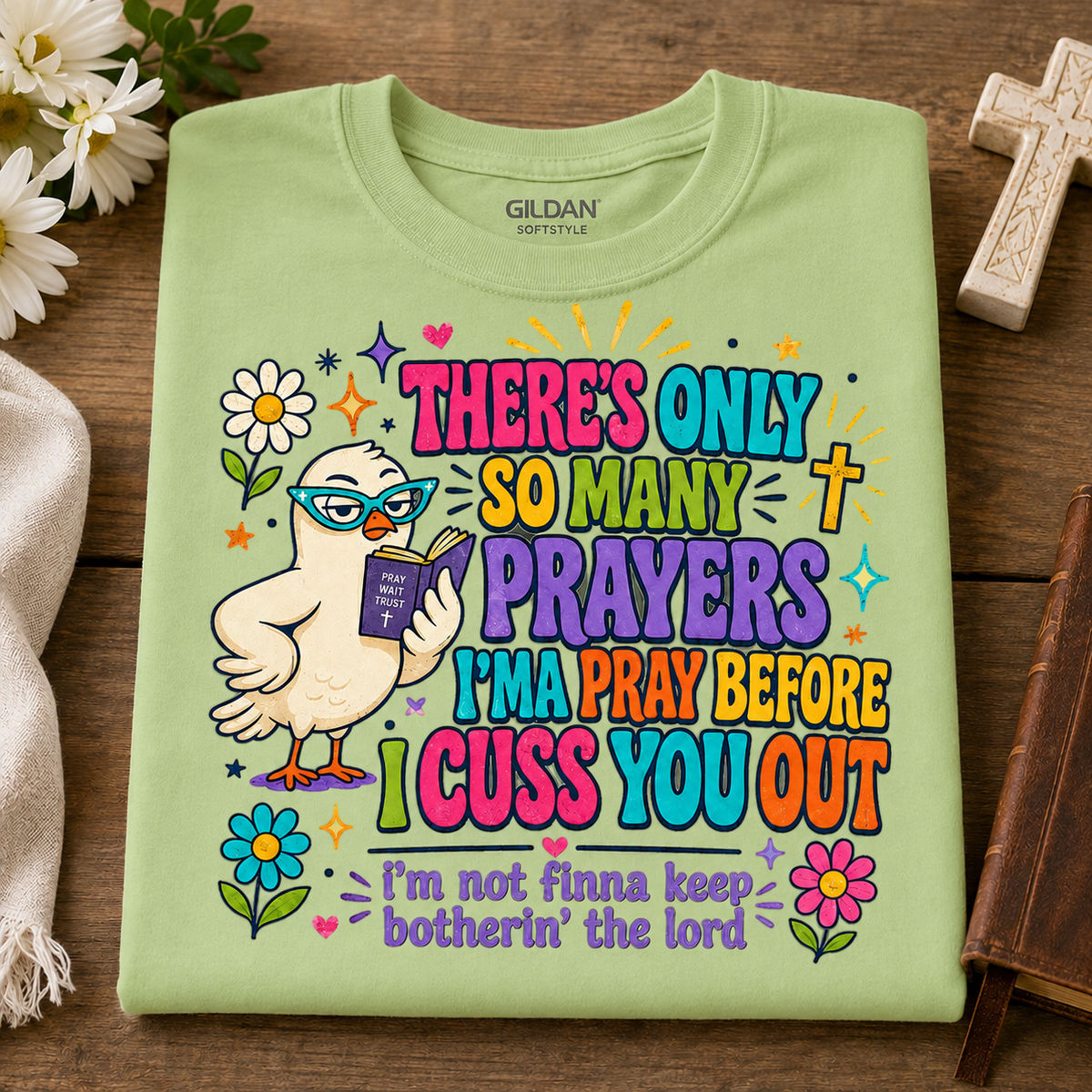 A folded pistachio colored Gildan Softstyle t-shirt with a colorful graphic of a cartoon bird wearing glasses and holding a Bible, surrounded by flowers and text that reads 'THERE'S ONLY SO MANY PRAYERS I'M A PRAY BEFORE I CUSS YOU OUT' and 'I'm not finna keep botherin' the lord'. The shirt is displayed flat on a wooden surface with decorative props.
