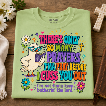 A folded pistachio colored Gildan Softstyle t-shirt with a colorful graphic of a cartoon bird wearing glasses and holding a Bible, surrounded by flowers and text that reads 'THERE'S ONLY SO MANY PRAYERS I'M A PRAY BEFORE I CUSS YOU OUT' and 'I'm not finna keep botherin' the lord'. The shirt is displayed flat on a wooden surface with decorative props.