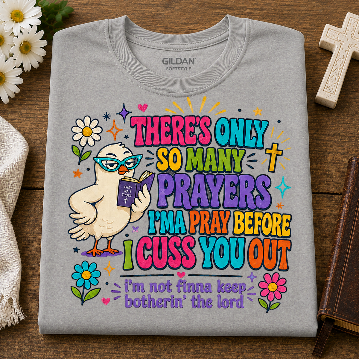 A folded sport grey Gildan t-shirt with a colorful graphic print featuring a cartoon duck wearing glasses and holding a Bible, surrounded by flowers, stars, and the text 'THERE'S ONLY SO MANY PRAYERS I'MA PRAY BEFORE I CUSS YOU OUT' and 'I'm not finna keep botherin' the lord'. The shirt is displayed on a wooden surface with decorative props including white daisies, a wooden cross, and a book.