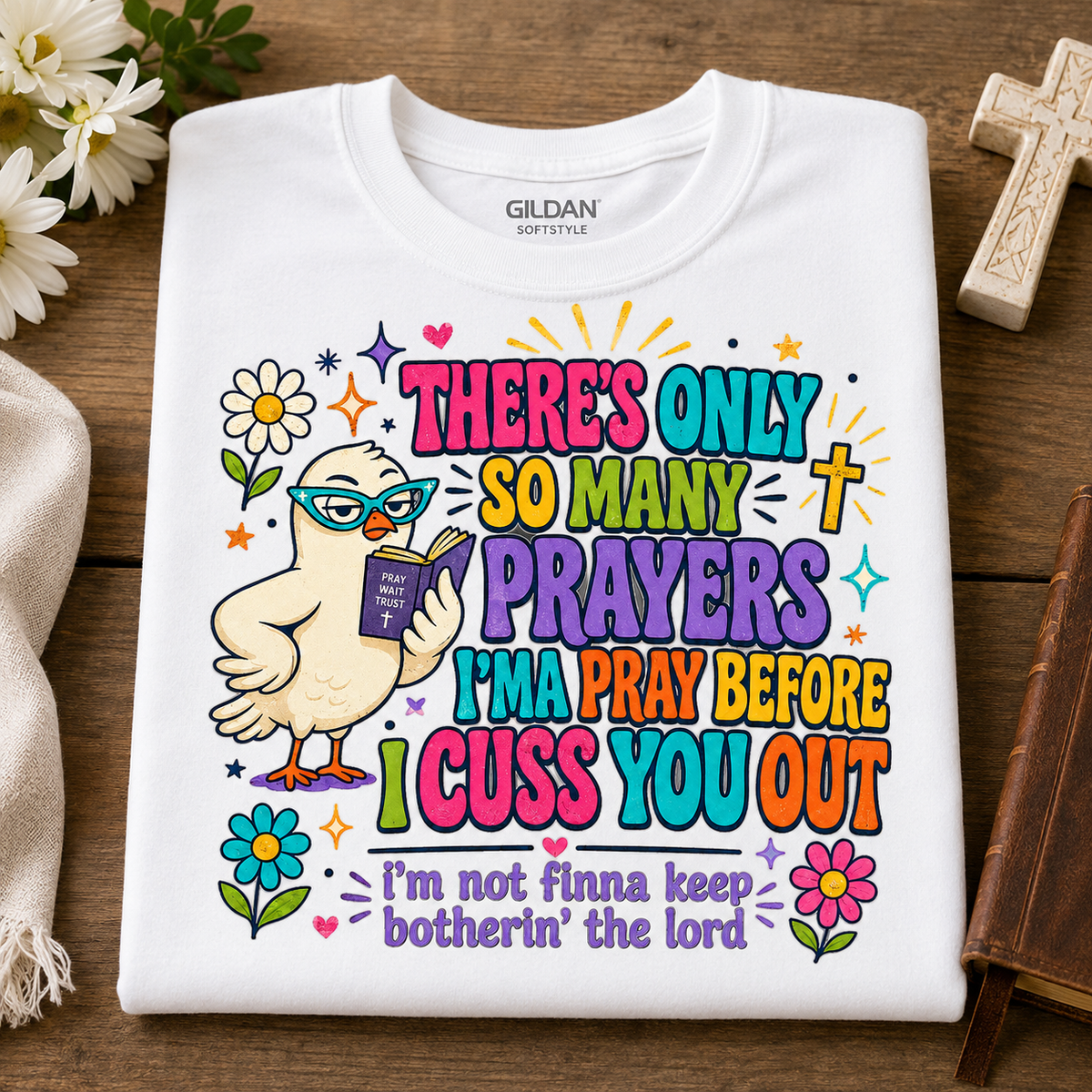 A folded white Gildan Softstyle t-shirt with a colorful graphic print, displayed on a wooden surface with decorative props including flowers, a cross, and a book.