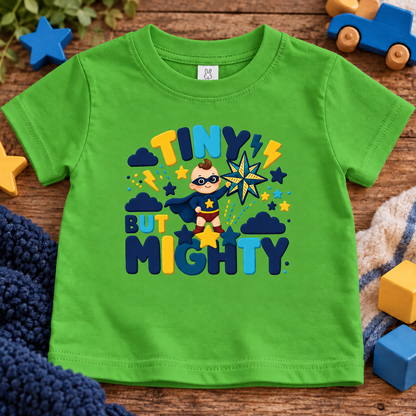 A apple green short-sleeve kids' t-shirt with a colorful 'TINY BUT MIGHTY' superhero graphic, laid flat on a wooden surface surrounded by toys.