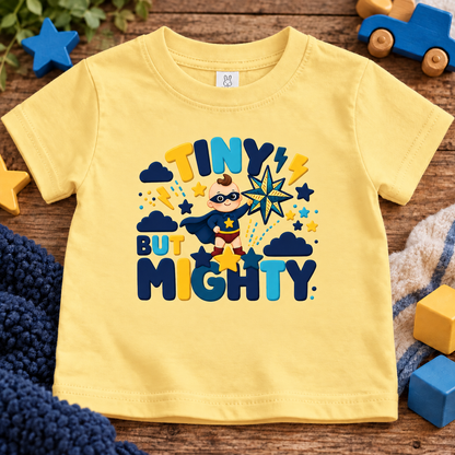 A butter short-sleeve baby t-shirt with a colorful graphic reading 'TINY BUT MIGHTY' and featuring a superhero character, laid flat on a wooden surface surrounded by toys and a knit blanket.