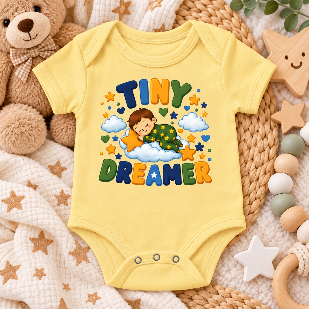 A butter yellow short-sleeve baby bodysuit laid flat on a cozy nursery surface. The front has a colorful cartoon graphic of a child sleeping on a cloud with stars and the words “TINY DREAMER” in large multicolored letters.