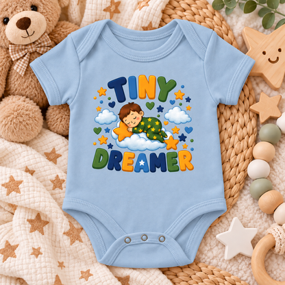 A light blue baby bodysuit with a colorful 'TINY DREAMER' graphic print, styled in a cozy flat-lay arrangement with a teddy bear, star toys, and a woven mat.