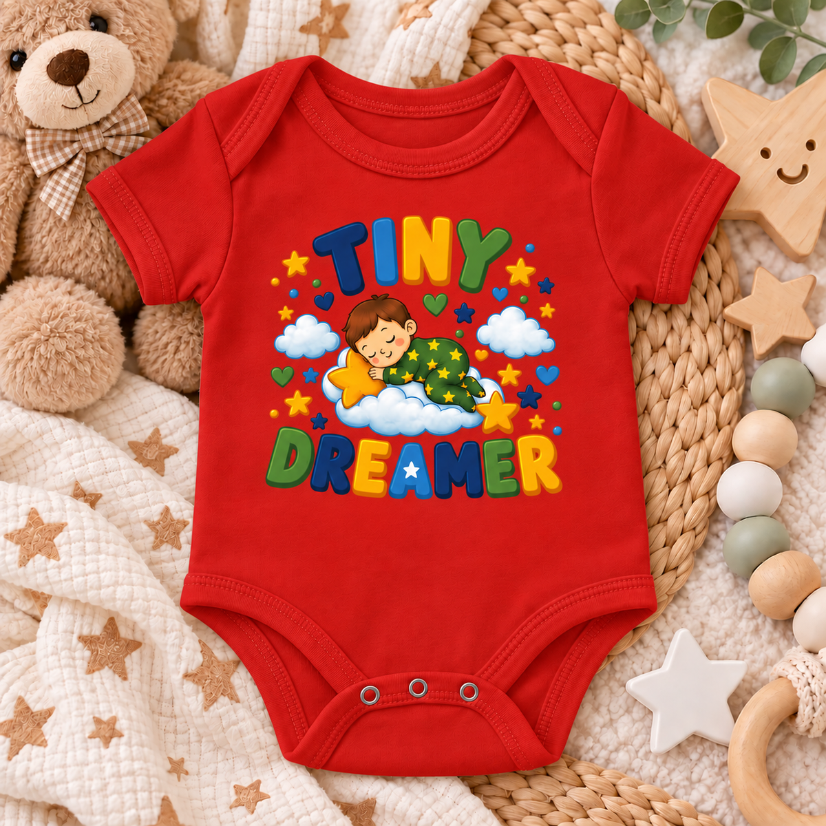 A red baby bodysuit with a colorful 'TINY DREAMER' graphic print, styled in a cozy flat-lay arrangement with a teddy bear, blanket, and wooden toys.