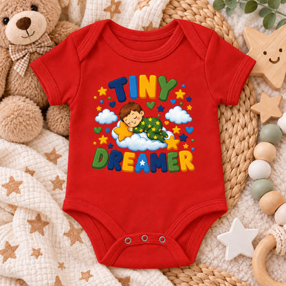 A red baby bodysuit with a colorful 'TINY DREAMER' graphic print, styled in a cozy flat-lay arrangement with a teddy bear, blanket, and wooden toys.