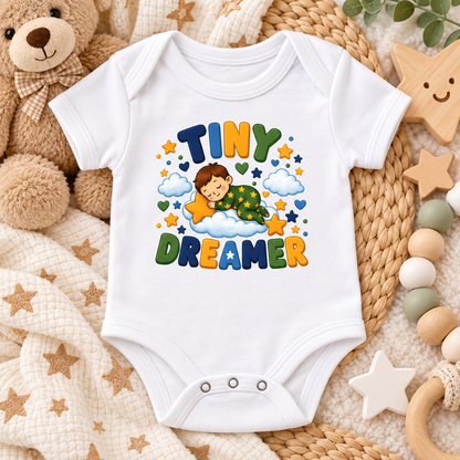 A white short-sleeve baby bodysuit laid flat on a cozy styled surface with a teddy bear, woven mat, wooden stars, beads, and a blanket. The bodysuit has a colorful printed graphic reading “TINY DREAMER” with a sleeping child, clouds, stars, and hearts.