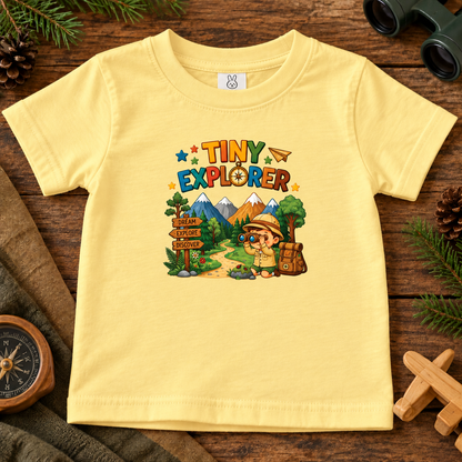 A butter yellow short-sleeve kids' t-shirt with a colorful 'TINY EXPLORER' graphic on the front, laid flat on a rustic wooden surface surrounded by outdoor-themed props like pine branches, a compass, binoculars, and a wooden cross.