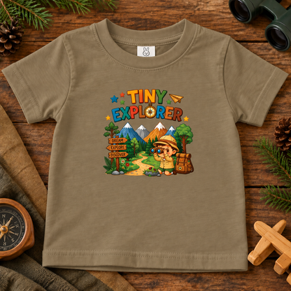 A toddler's short-sleeve t-shirt in a warm coyote brown color, featuring a colorful cartoon graphic of a child explorer standing in front of mountains with the text 'TINY EXPLORER'. The shirt is displayed in a flat-lay setting on a rustic wooden surface surrounded by outdoor-themed props like pine branches, a compass, binoculars, and a wooden airplane.