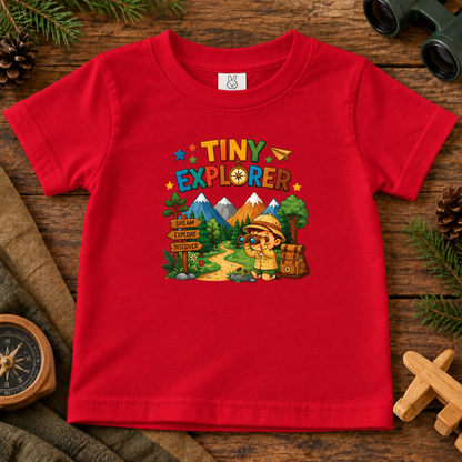 A red short-sleeve kids' t-shirt with a colorful 'TINY EXPLORER' graphic on the chest, displayed in a flat-lay arrangement on a rustic wooden surface with outdoor-themed props like pine branches, a compass, and binoculars.