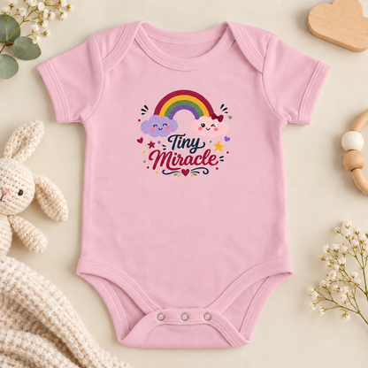 A pink short-sleeve baby bodysuit laid flat on a cream background, decorated with a colorful cartoon rainbow, clouds, and the words 'Tiny Miracle' in script. The bodysuit is styled with baby props including a plush bunny, dried flowers, and wooden toys.