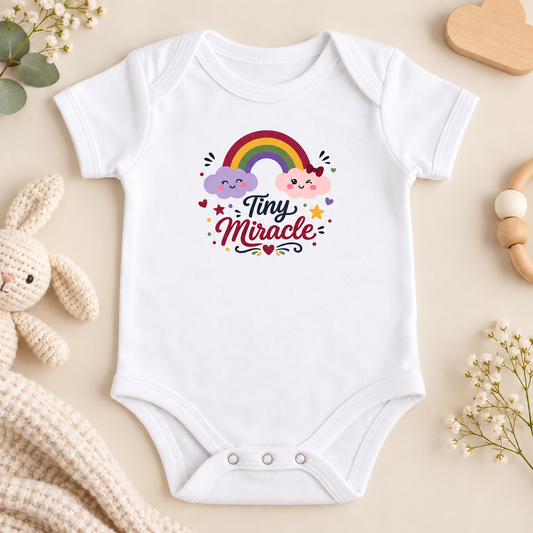 A white onesie(bodysuit) with a rainbow and clouds graphic with text Tiny Miracle