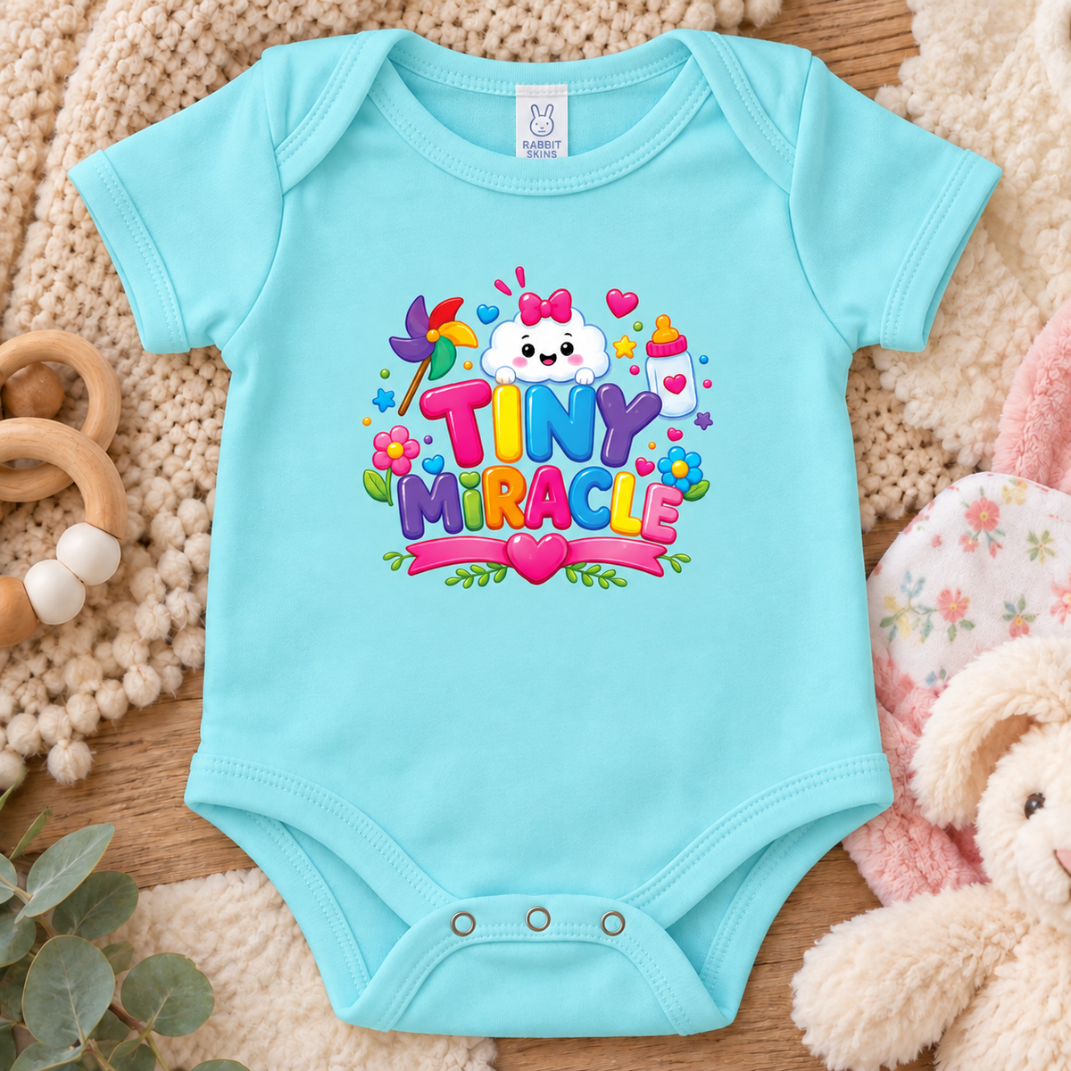 A chlll blue colored short-sleeve baby bodysuit features a colorful 'TINY MIRACLE' graphic with a cartoon cloud, flowers, hearts, and a baby bottle.