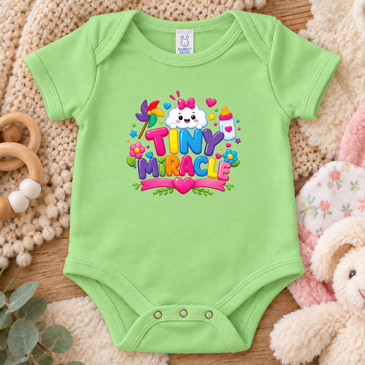 A keylime green short-sleeve baby bodysuit features a colorful 'TINY MIRACLE' graphic with a cartoon cloud, flowers, hearts, and a baby bottle.