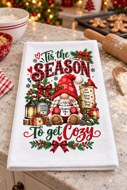 A white kitchen towel with a festive Christmas gnome design and the text 'Tis the Season To get Cozy' is displayed on a kitchen counter surrounded by holiday baking props.