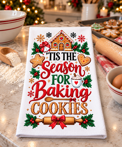 A white kitchen towel with a festive Christmas baking design, featuring the text 'Tis the Season for Baking Cookies' in red and gold lettering, surrounded by gingerbread houses, cookies, holly, and a rolling pin. The towel is displayed on a marble countertop with baking ingredients and a blurred holiday background.