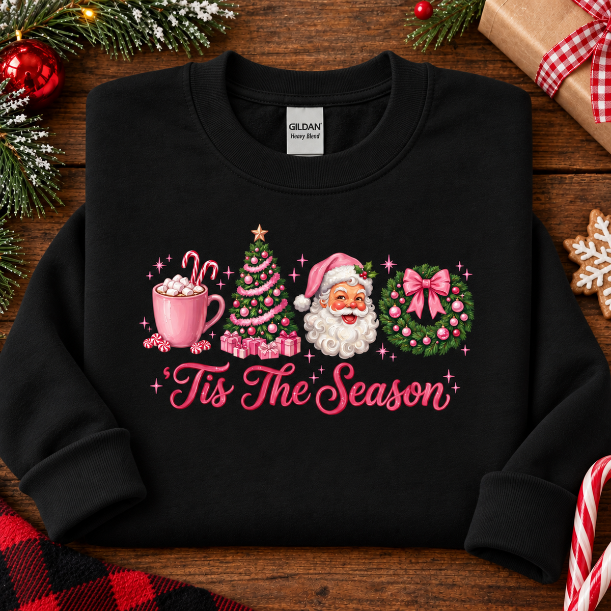 A black crewneck sweatshirt with a colorful Christmas graphic on the front, laid flat on a rustic wooden background decorated with holiday ornaments.The graphic features a pink mug, a Christmas tree, Santa Claus, a wreath, and the text 'Tis The Season'.