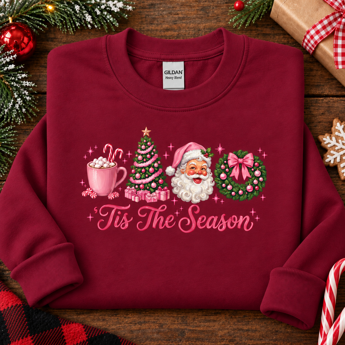 A folded cardinal red crewneck sweatshirt with a colorful Christmas graphic and the text 'Tis The Season' on the front. The graphic includes a mug of hot cocoa, a Christmas tree, Santa Claus, and a wreath.