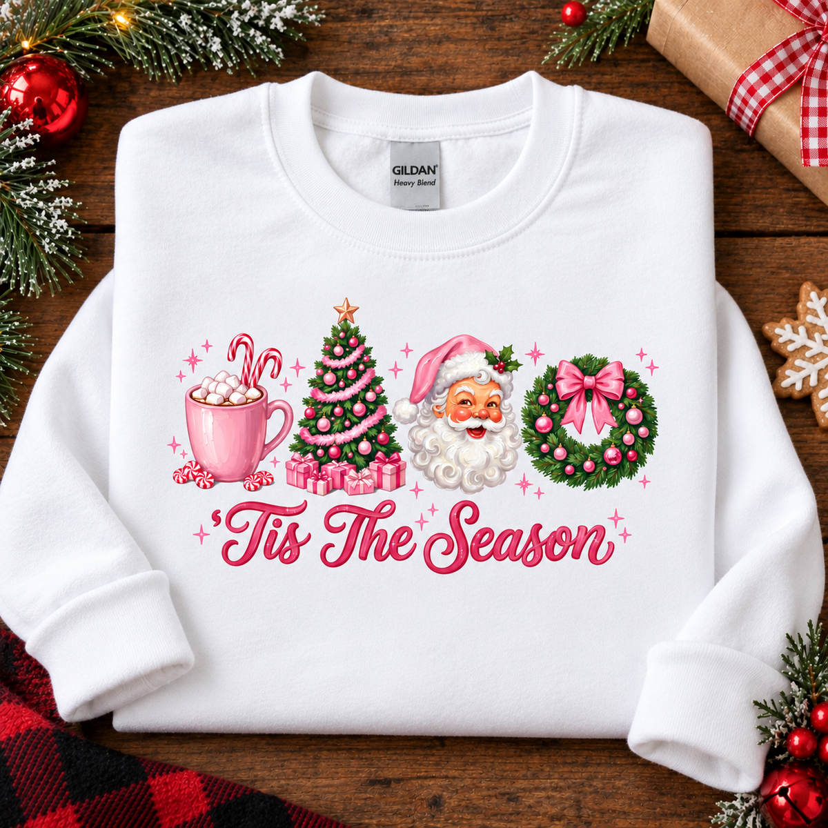 A white long-sleeve crewneck sweatshirt laid flat on a wooden surface, decorated with a colorful Christmas graphic that reads 'Tis The Season.' The design includes a pink mug with hot cocoa and marshmallows, a green Christmas tree, Santa Claus, and a green wreath with pink accents.