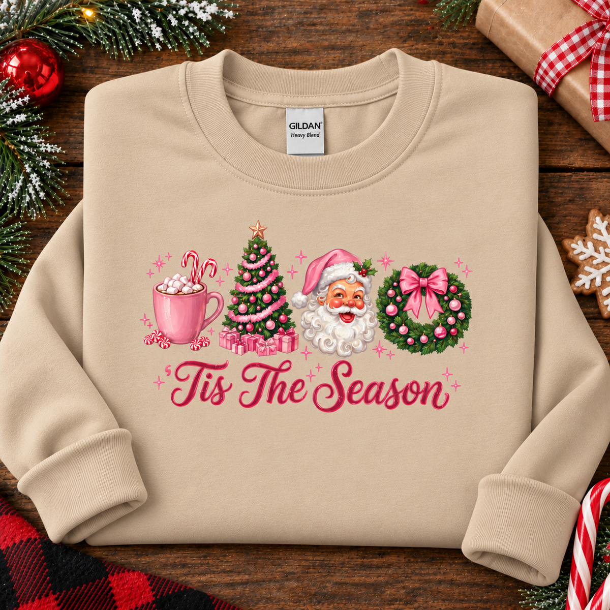 A sand colored crewneck sweatshirt laid flat on a rustic wooden surface, decorated with Christmas-themed graphics and the text 'Tis The Season.' The print shows a pink mug with candy canes, a green Christmas tree, Santa Claus, and a pink bow on a wreath.